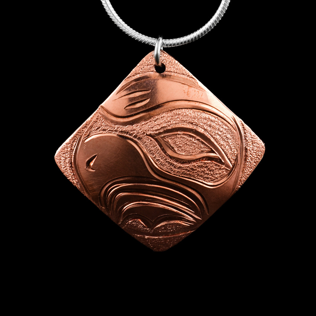 This Eagle pendant has a square, diamond shaped and is made from copper. The head of the Eagle is carved in a side profile in the center of the piece. The eye, beak and feather designs are detailed and there are dimpled designs in the negative spaces of the scene.