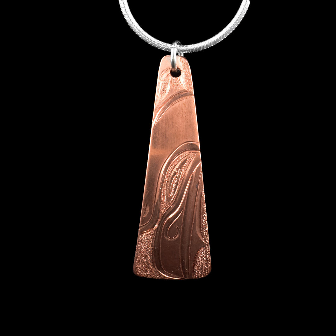 This Raven pendant has a long, thin and oblong shape that tapers out at the bottom. The piece is made from copper and the head of the Raven is carved into the it at a side profile. The eye, beak and feather designs are all detailed. There are dimpled textures in the negative spaces of the scene.
