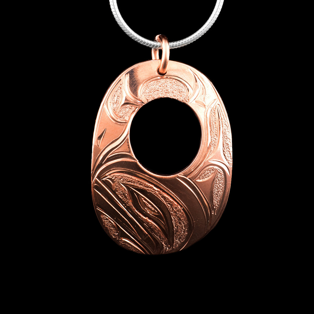 This Orca pendant was a wide, oval shape and a smaller, circle cutout in the center of it. The piece is made from copper and there is a depiction of the Orca legend carved into the surface of it. The head of the Orca is at the bottom of the piece and the rest of its body spans around the of it with its dorsal fin being on the right side above the head. The head is done in a side profile and there are dimpled textures in the negative spaces of the scene.