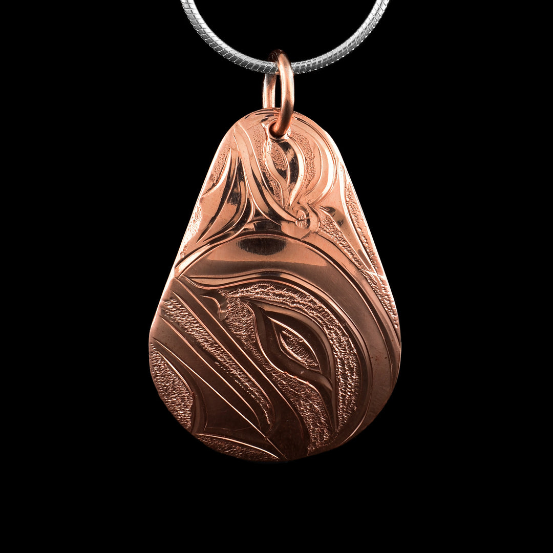 This Orca pendant has a ide, teardrop shape and is made from copper. The head of the Orca is carved into the surface of the piece at a side profile. It is looking up wards and there are swirling designs on the top of the piece. There are small, dimpled textures in the negative space of the scene.