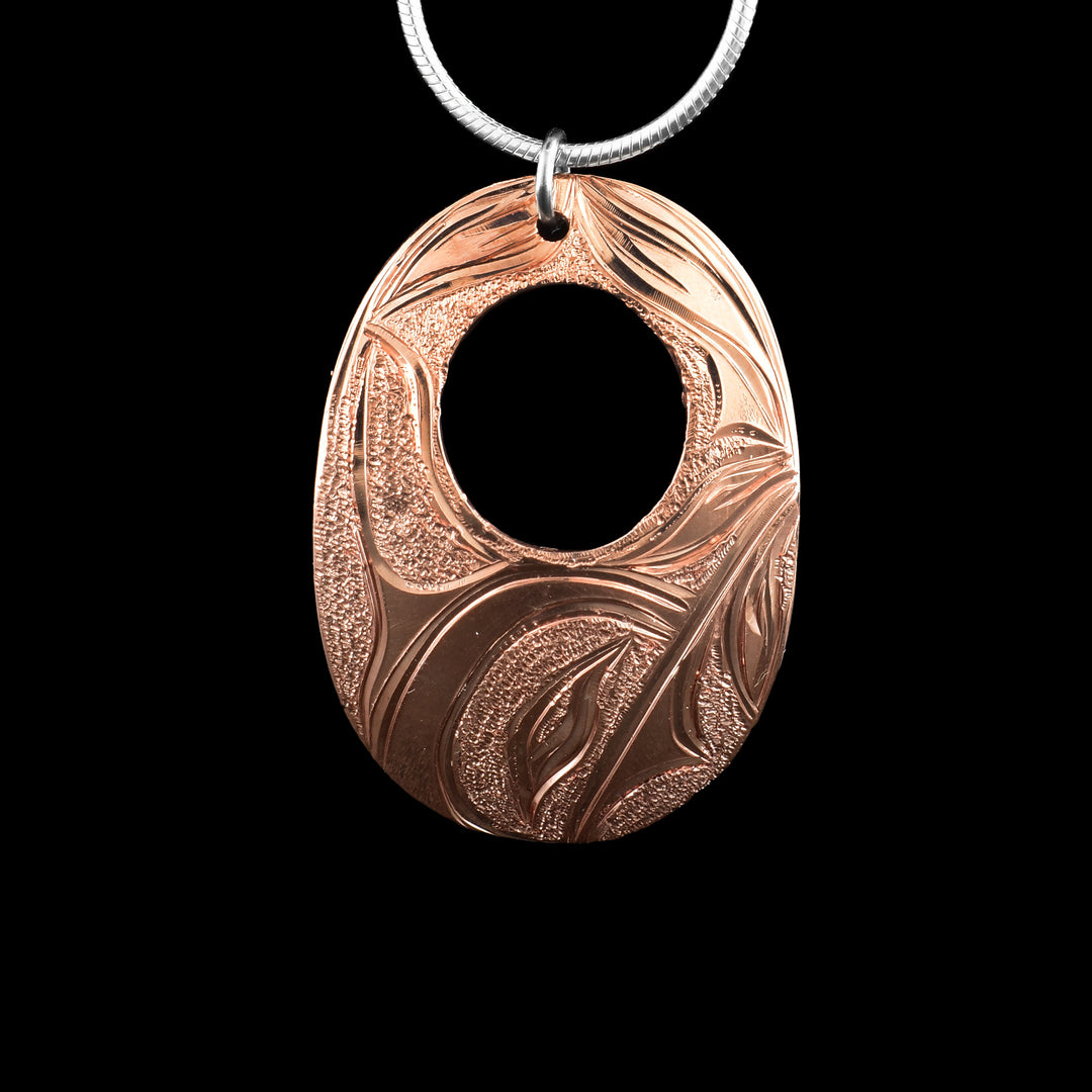 This Hummingbird pendant was a wide, oval shape and a smaller, circle cutout in the center of it. The piece is made from copper and there is a depiction of the Hummingbird legend carved into the surface of it. The head of the Hummingbird is at the bottom of the piece and its wings extend up and around the rest of the piece. There are dimpled textures in the negative spaces of the scene.