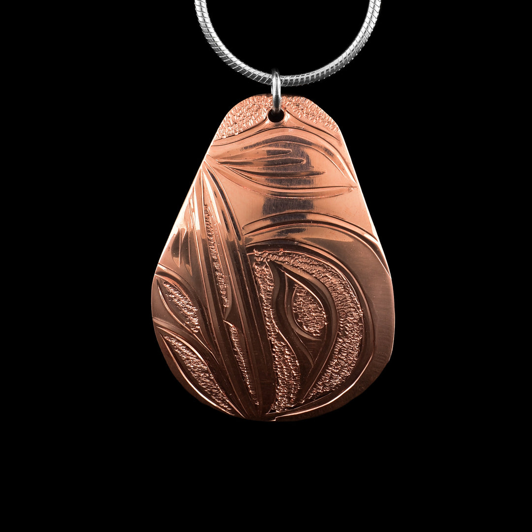 This Hummingbird pendant has a wide, teardrop shape and is made from copper. The head of the Hummingbird is carved at a side profile in the center of the piece. Its beak is facing upwards and there are two flower petal designs at the top where the legends beak is. There are dimpled textures in the negative spaces of the scene.