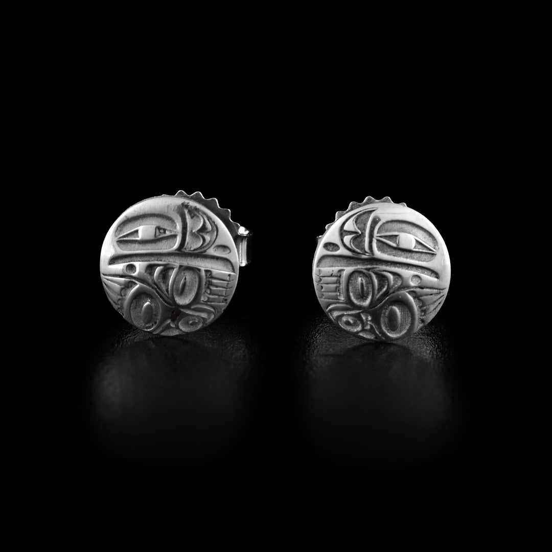 These Raven stud earrings are made from sterling silver and have a small, circle shape. There is an abstract depiction of the Raven made in the surface of the studs. The eye and beak is at the top and there are two wings on top of each other at the bottom.