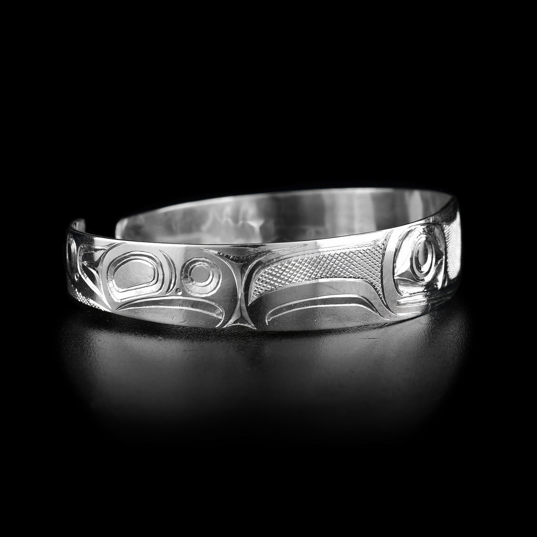 This frog cuff bracelet has a single band with a gap in the back made from sterling silver. They Frog legend is depicted across the band with its two, large eyes being at the front of the piece. Its mouth is shown below the eyes as well as its nose. There are designs representing its legs across the rest of the band. There are cross hatching designs in the negative spaces of the scene.