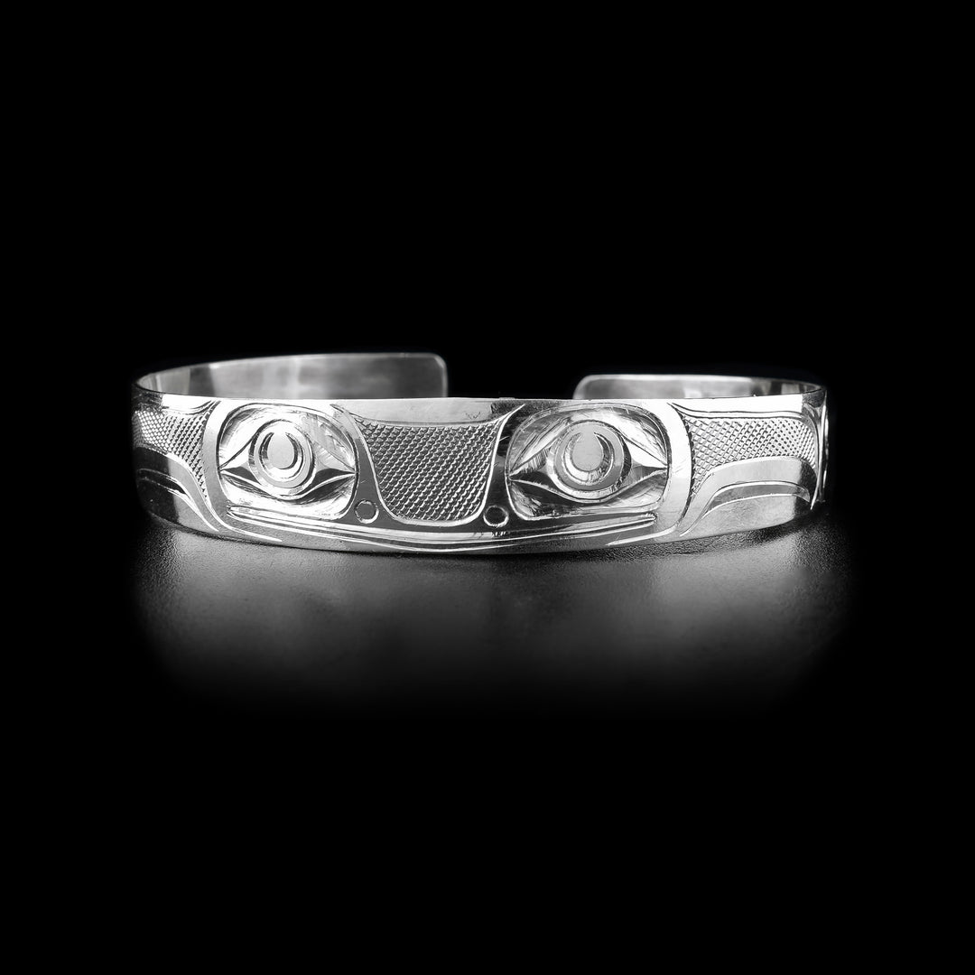 This frog cuff bracelet has a single band with a gap in the back made from sterling silver. They Frog legend is depicted across the band with its two, large eyes being at the front of the piece. Its mouth is shown below the eyes as well as its nose. There are designs representing its legs across the rest of the band. There are cross hatching designs in the negative spaces of the scene.