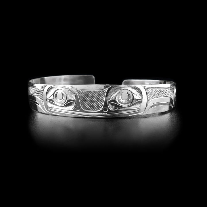 This frog cuff bracelet has a single band with a gap in the back made from sterling silver. They Frog legend is depicted across the band with its two, large eyes being at the front of the piece. Its mouth is shown below the eyes as well as its nose. There are designs representing its legs across the rest of the band. There are cross hatching designs in the negative spaces of the scene.