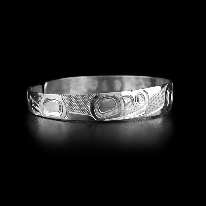This Hummingbird cuff bracelet has a single, thin band made from sterling silver with a gap in the back. There is a depiction of the Hummingbird made into the band with its head in the front shown at a side profile. Its beak extends across the band and at the end of it, there is a flower. There are indigenous style designs at the ends of the band and cross hatching patterns in the negative spaces of the scene.