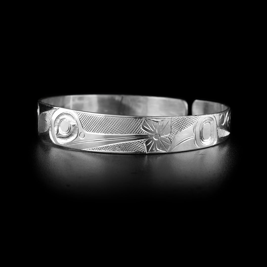 This Hummingbird cuff bracelet has a single, thin band made from sterling silver with a gap in the back. There is a depiction of the Hummingbird made into the band with its head in the front shown at a side profile. Its beak extends across the band and at the end of it, there is a flower. There are indigenous style designs at the ends of the band and cross hatching patterns in the negative spaces of the scene.