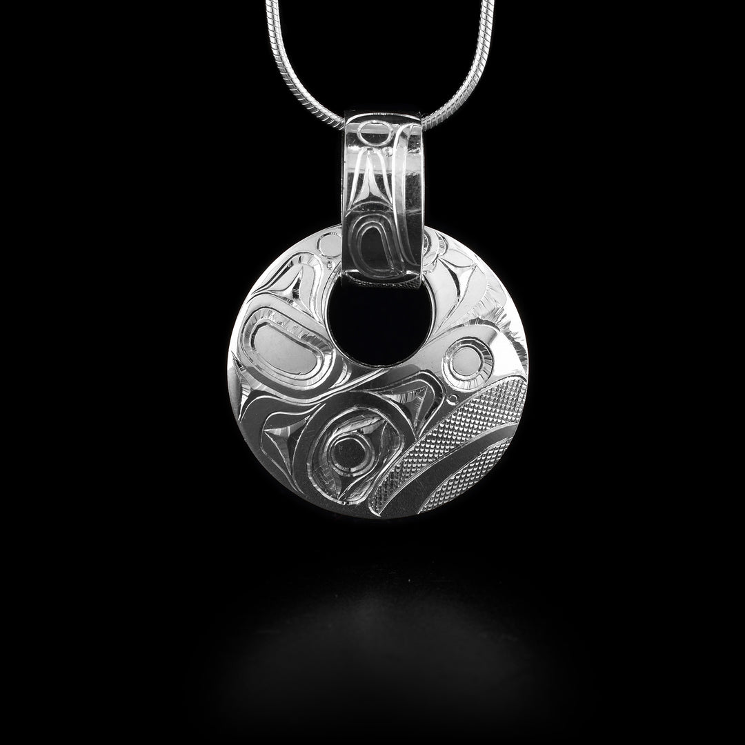 This sterling silver Eagle pendant has a circle shape with a small, circle cutout close to the top of the piece. There is a wide, decorative bail linked through the cutout with a depiction of the Eagle and designs carved into it. The head of the Eagle is depicted at a side profile at the bottom of the pendant and designs representing its wings across the rest.