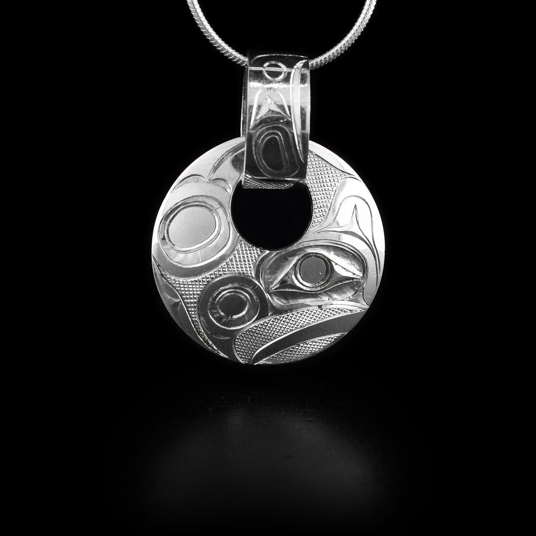 This sterling silver Raven pendant has a circle shape with a small, circle cutout close to the top of the piece. There is a wide, decorative bail linked through the cutout with a depiction of the Raven and designs carved into it. The head of the Raven is depicted at a side profile at the bottom of the pendant and designs representing its wings across the rest.