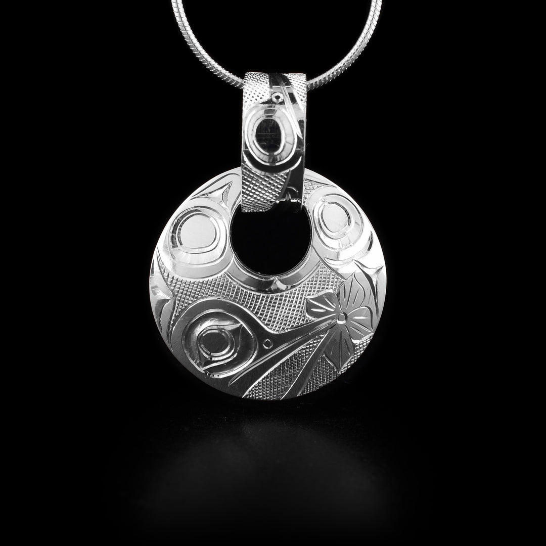 This sterling silver Hummingbird pendant has a circle shape with a small, circle cutout close to the top of the piece. There is a wide, decorative bail linked through the cutout with a depiction of the Hummingbird and designs carved into it. The head of the Hummingbird is depicted at a side profile at the bottom of the pendant with a flower at the end of its beak. There are designs representing its wings across the rest.
