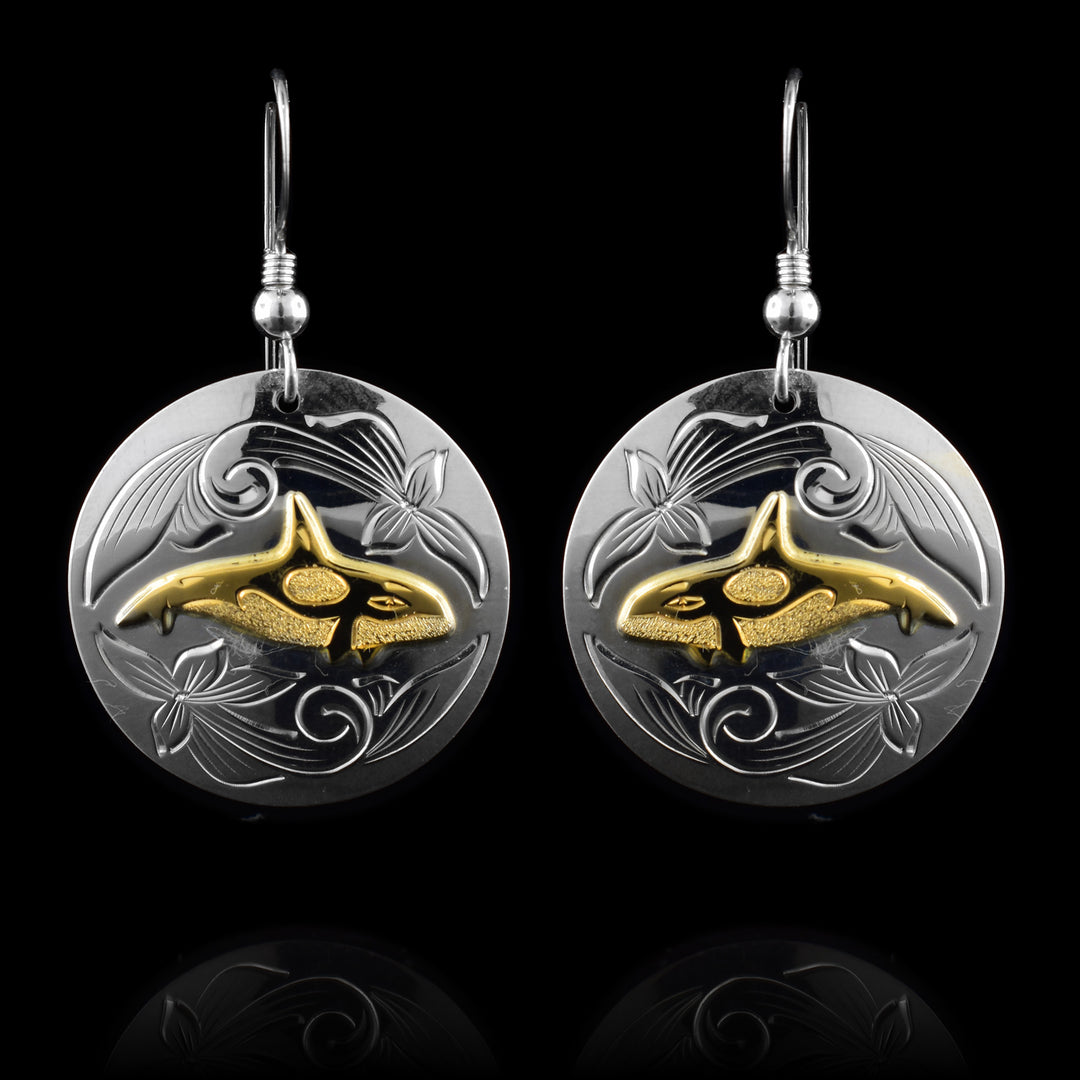 These sterling silver and 14K gold Orca whale earrings have circle shaped hangs. There is a side profile of the Orca's body made with the gold in the center of the hangs. Its white spots are highlighted with dimpled spots. There are swirling designs carved into the silver around the Orca.