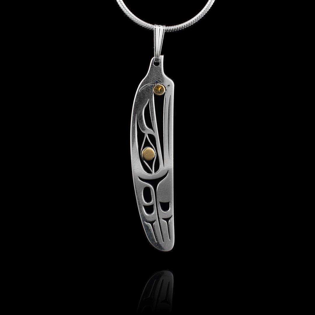 This Raven feather pendant has been made from sterling silver and 14K gold. The pendant is mostly silver and is shaped like a feather. There are cutouts on the piece depicting the Raven legend with its head at the top facing up and a feather, wing- like design behind the head. The eye and pearl held in its beak are done in gold.