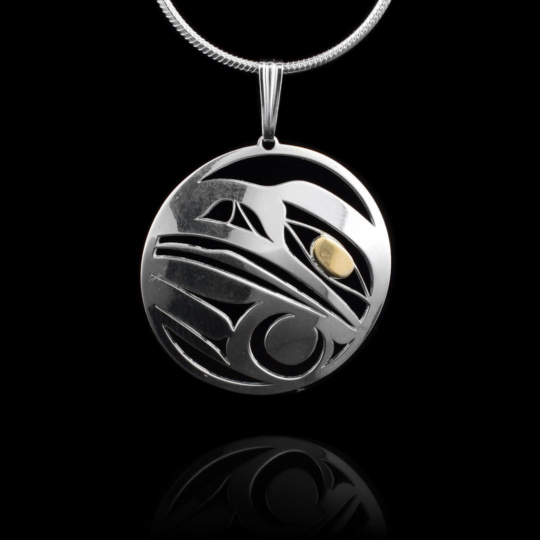 This sterling silver and 14K gold Raven pendant is circle shaped and there are cutout designs depicting the Raven. The head of the Raven is shown at a side profile and there is a feather design below the head that represents a wing. The eye is done using the gold.