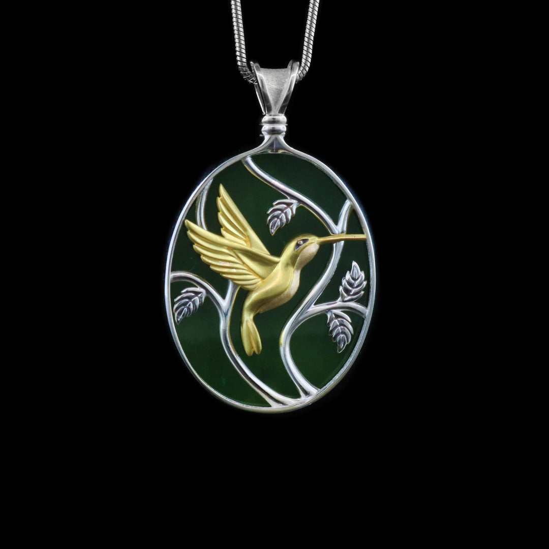 This jade pendant necklace has an oval shaped with a depiction of a Hummingbird flying around a tree. The hummingbird is made from 14K gold and the jade and three designs are set in sterling silver.