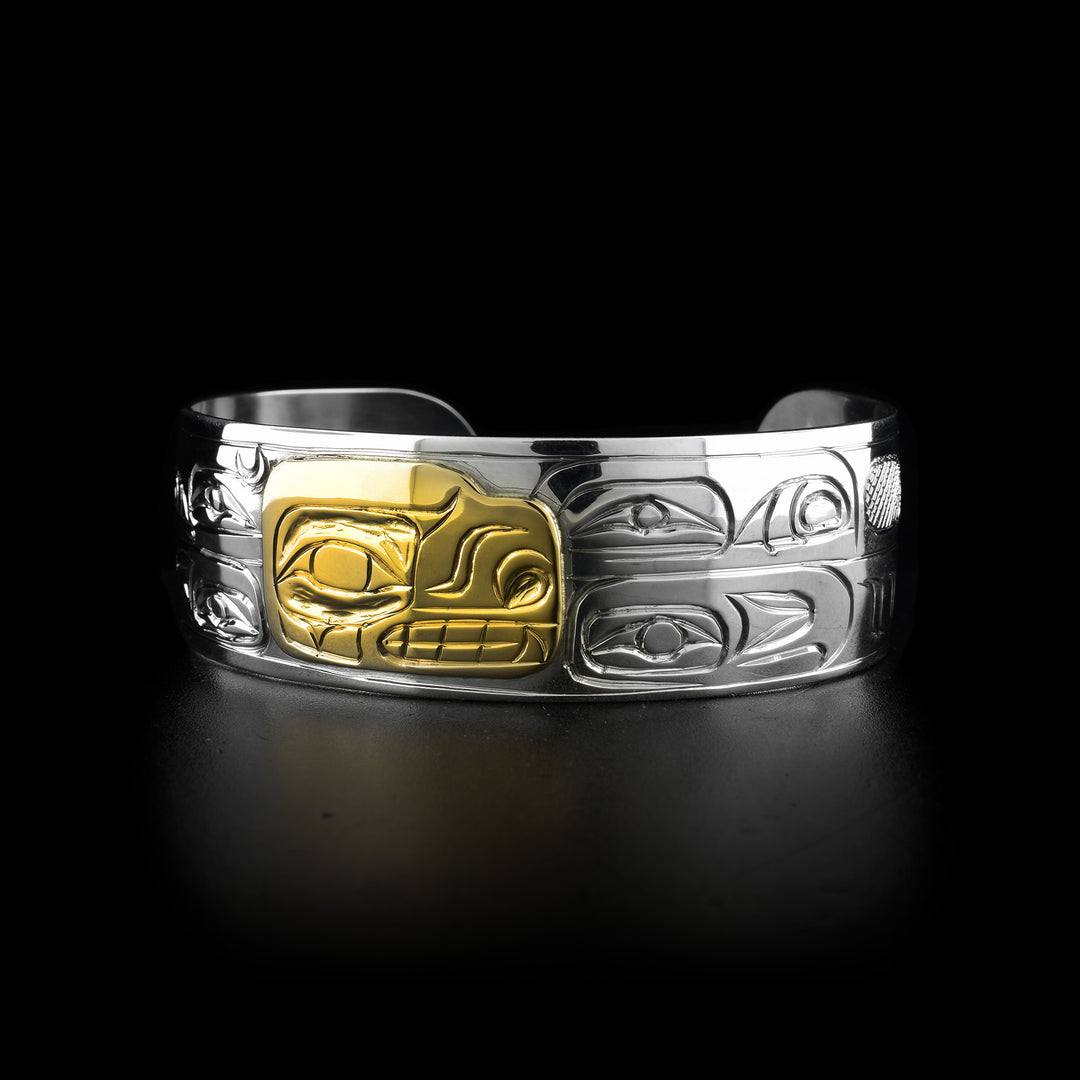 This gold Bear bracelet has a single, wide band with a gap in the back. The band is made from sterling silver and there is a depiction of the head of the Bear done at a side profile in 14K gold. There are unique carvings made around the rest of the band depicting eyes.