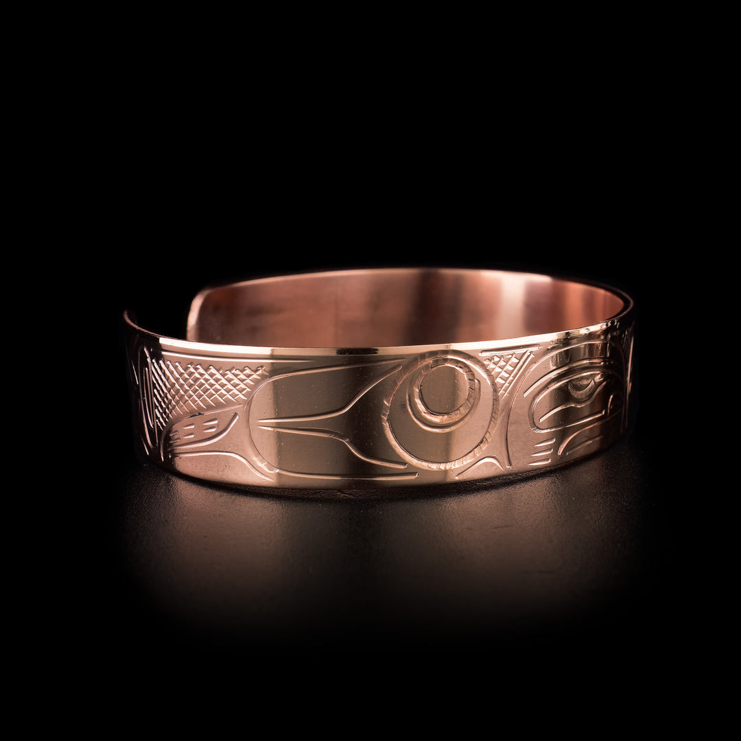 This copper cuff bracelet has a single band with a gap in the back and two depictions of the Orca carved into it. The heads of the Orcas meet at the top of the band and are facing each other. There are designs representing their dorsal fins and body behind the heads and cross hatching patterns in the negative spaces of the scene.