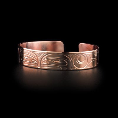 This copper cuff bracelet has a single band with a gap in the back and two depictions of the Orca carved into it. The heads of the Orcas meet at the top of the band and are facing each other. There are designs representing their dorsal fins and body behind the heads and cross hatching patterns in the negative spaces of the scene.