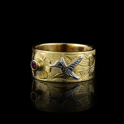 This ruby band ring gold has a wide band made from 10K gold. There are floral designs carved into the band and one of the flowers has a ruby in the center of it. There is a hummingbird done in sterling silver with its beak facing towards the ruby.
