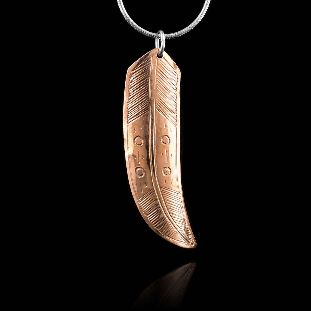 This copper feather pendant is long and curves out at the bottom. The piece is made form copper and shaped like a feather. There are intricate feather designs carved into the surface of the piece.