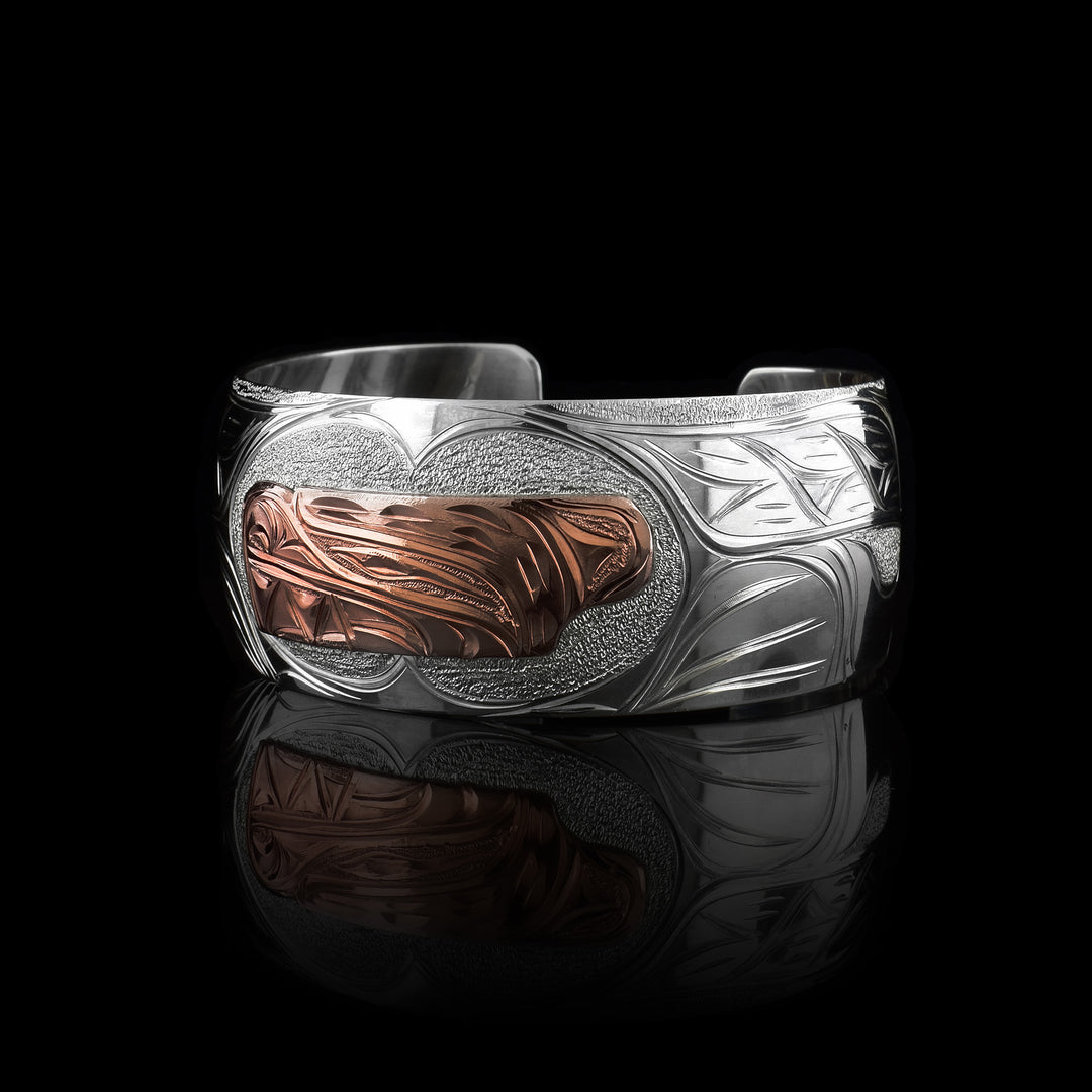 This sterling silver and copper bracelet has a single, wide band with a gap in the back. The head of the Bear is depicted at a side profile using copper and is in the center of the band. There are claw and paw designs made on either side of the Bear head and has a tail detail on one end.
