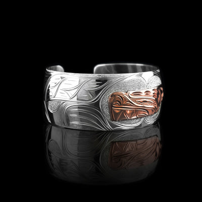 This sterling silver and copper bracelet has a single, wide band with a gap in the back. The head of the Bear is depicted at a side profile using copper and is in the center of the band. There are claw and paw designs made on either side of the Bear head and has a tail detail on one end.