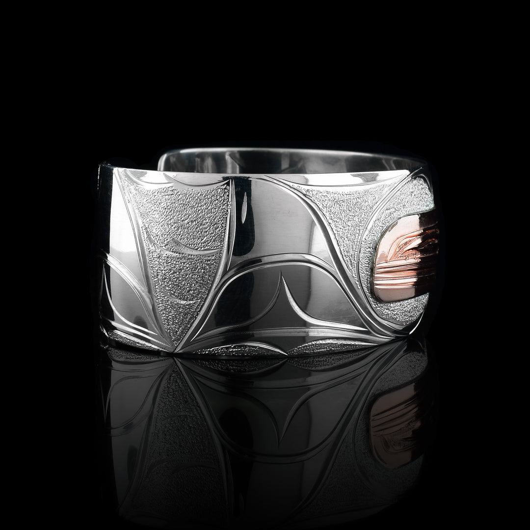 This sterling silver and copper bracelet has a single, wide band with a gap in the back. The head of the Orca is depicted t a side profile using copper and is in the center of the band. There are wave, like designs around the Orca head as well as a dorsal fin.