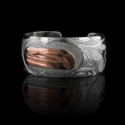 This sterling silver and copper bracelet has a single, wide band with a gap in the back. The head of the Orca is depicted t a side profile using copper and is in the center of the band. There are wave, like designs around the Orca head as well as a dorsal fin.