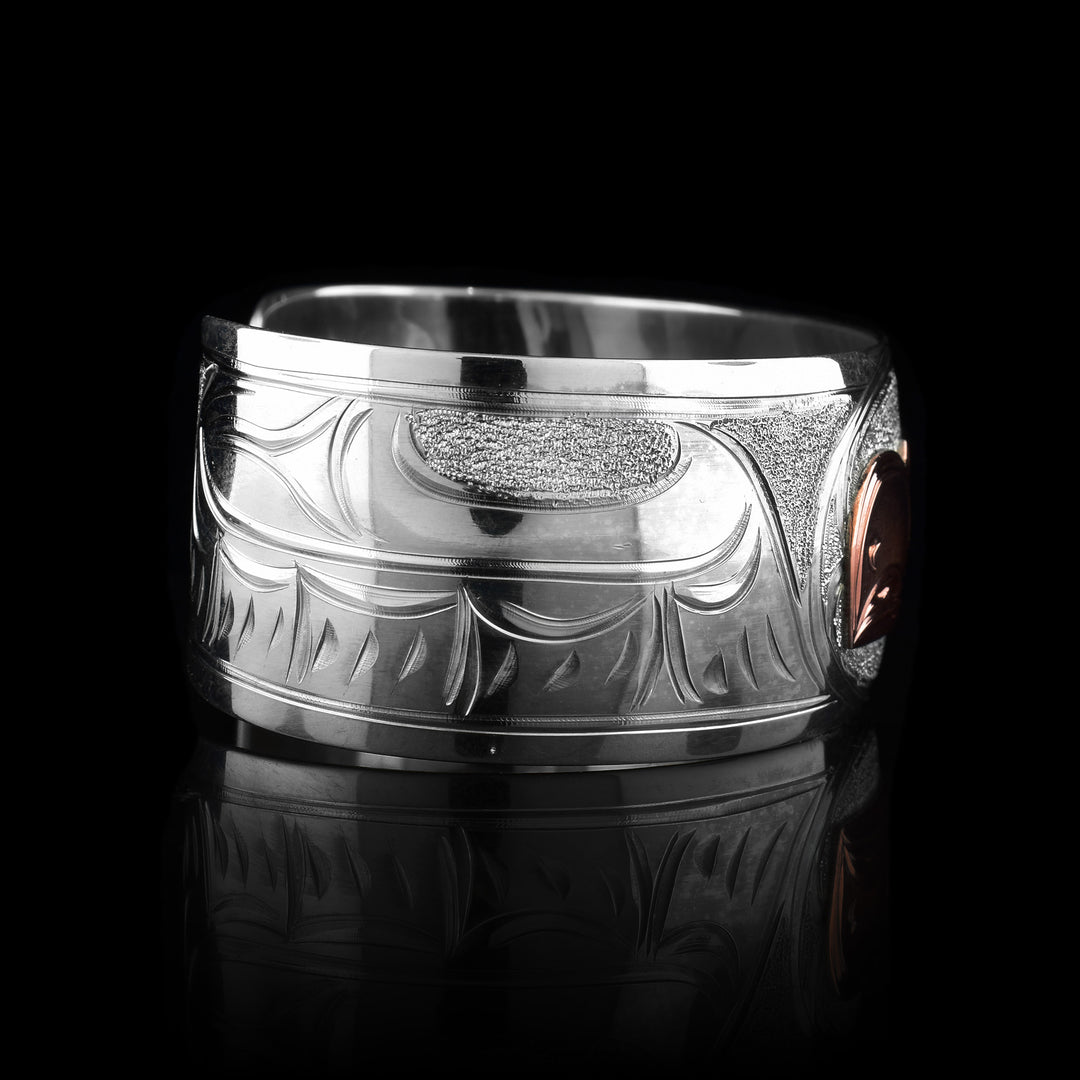 This sterling silver copper bracelet has a single, wide band with a gap in the back and the head of the Eagle is depicted in the center of it. The head is done in copper and at a side profile. On both side of the band, there are carvings showing the wings outstretched with feather details.
