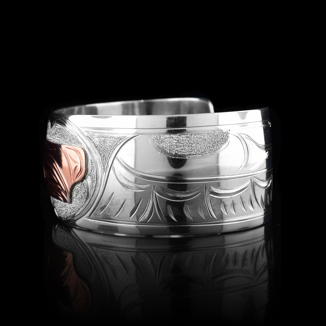 This sterling silver copper bracelet has a single, wide band with a gap in the back and the head of the Eagle is depicted in the center of it. The head is done in copper and at a side profile. On both side of the band, there are carvings showing the wings outstretched with feather details.
