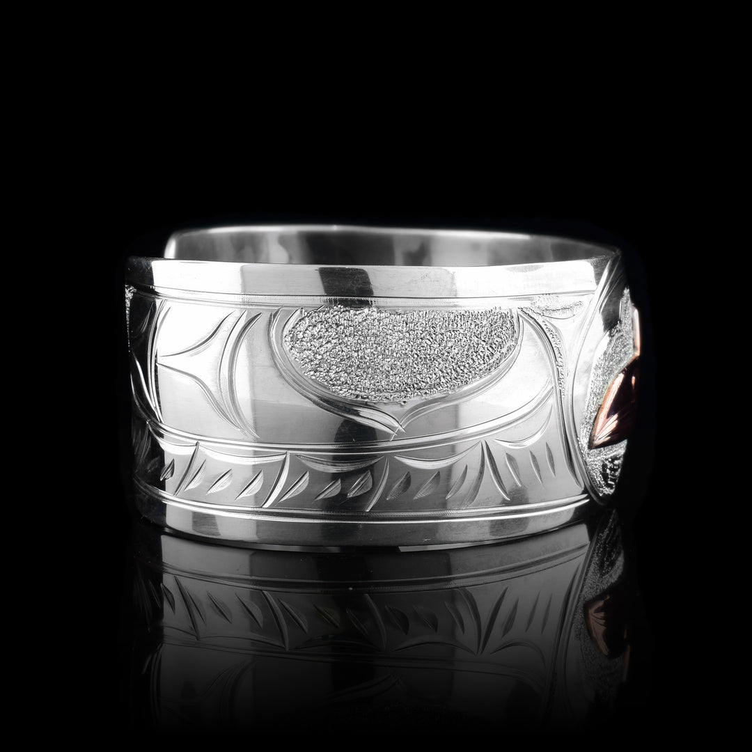 This sterling silver copper bracelet has a single, wide band with a gap in the back and the head of the raven is depicted in the center of it. The head is done in copper and at a side profile. On both side of the band, there are carvings showing the wings outstretched across the band with feather details.