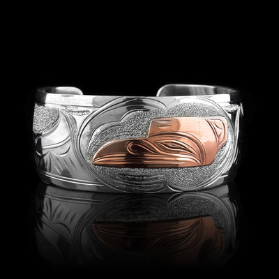 This sterling silver copper bracelet has a single, wide band with a gap in the back and the head of the raven is depicted in the center of it. The head is done in copper and at a side profile. On both side of the band, there are carvings showing the wings outstretched across the band with feather details.
