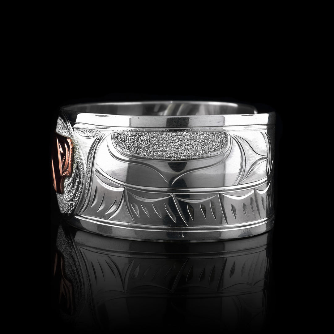 This sterling silver copper bracelet has a single, wide band with a gap in the back and the head of the raven is depicted in the center of it. The head is done in copper and at a side profile. On both side of the band, there are carvings showing the wings outstretched across the band with feather details.