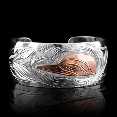 This large sterling silver copper bracelet has a single, wide band with a gap in the back. The head of the Hummingbird is done at a side profile using copper and there is a wing designs carved behind it on the band. On the other side there are floral design and its beak is reaching between two flower petals.