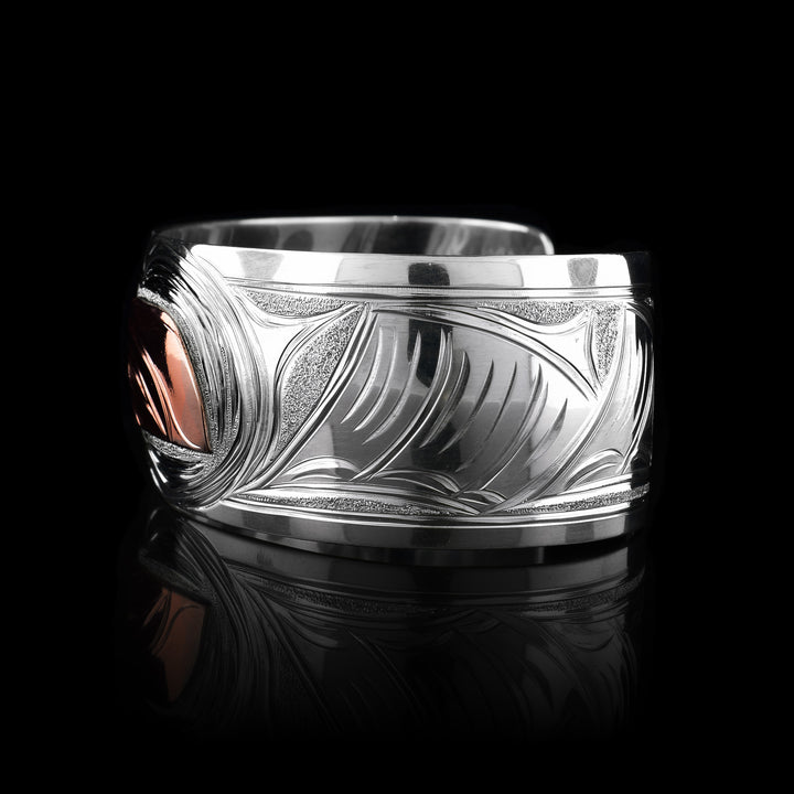 This large sterling silver copper bracelet has a single, wide band with a gap in the back. The head of the Hummingbird is done at a side profile using copper and there is a wing designs carved behind it on the band. On the other side there are floral design and its beak is reaching between two flower petals.