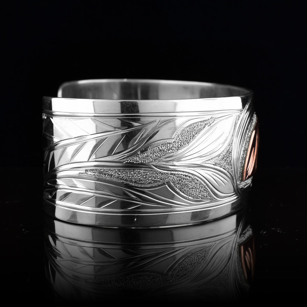 This Hummingbird bracelet with moon has a single, wide band made from sterling silver. The moon legend is depicted as a half circle cut horizontally and is done using copper. You can see half of the face of the Moon legend. On one end of the band, there are floral designs in silver including around the Moon. On the other side, there is a Hummingbird head done at a side profile with its beak reaching into the moon.