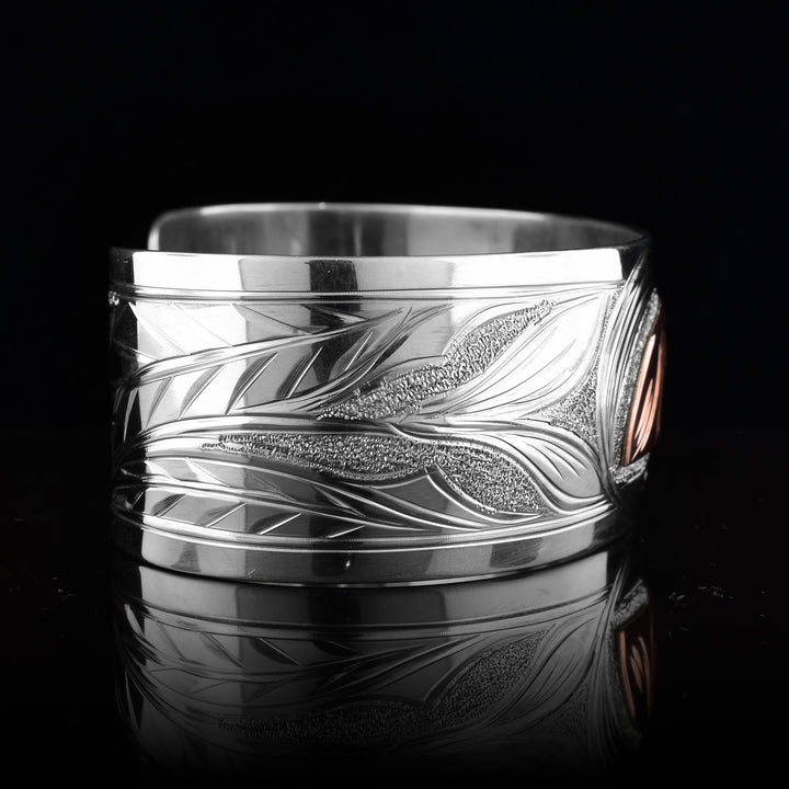 This Hummingbird bracelet with moon has a single, wide band made from sterling silver. The moon legend is depicted as a half circle cut horizontally and is done using copper. You can see half of the face of the Moon legend. On one end of the band, there are floral designs in silver including around the Moon. On the other side, there is a Hummingbird head done at a side profile with its beak reaching into the moon.