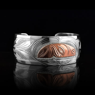 This Hummingbird bracelet with moon has a single, wide band made from sterling silver. The moon legend is depicted as a half circle cut horizontally and is done using copper. You can see half of the face of the Moon legend. On one end of the band, there are floral designs in silver including around the Moon. On the other side, there is a Hummingbird head done at a side profile with its beak reaching into the moon.