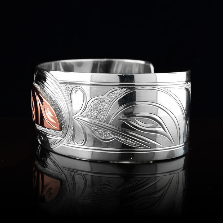 This Hummingbird bracelet with moon has a single, wide band made from sterling silver. The moon legend is depicted as a half circle cut horizontally and is done using copper. You can see half of the face of the Moon legend. On one end of the band, there are floral designs in silver including around the Moon. On the other side, there is a Hummingbird head done at a side profile with its beak reaching into the moon.