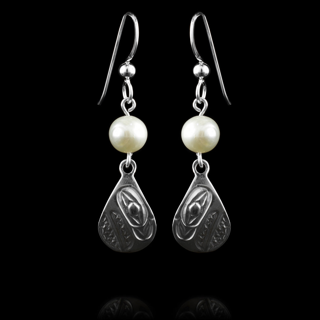 These dangling teardrop earrings have teardrop shaped hangs cast from sterling silver. There is a single, white pearl below the hooks and above the hangs. The head of the Raven is cast into the hangs at a side profile with its beak facing down. The beak and eye are large and takes up most of the hangs. There are cross hatching designs in the background of the portrait.