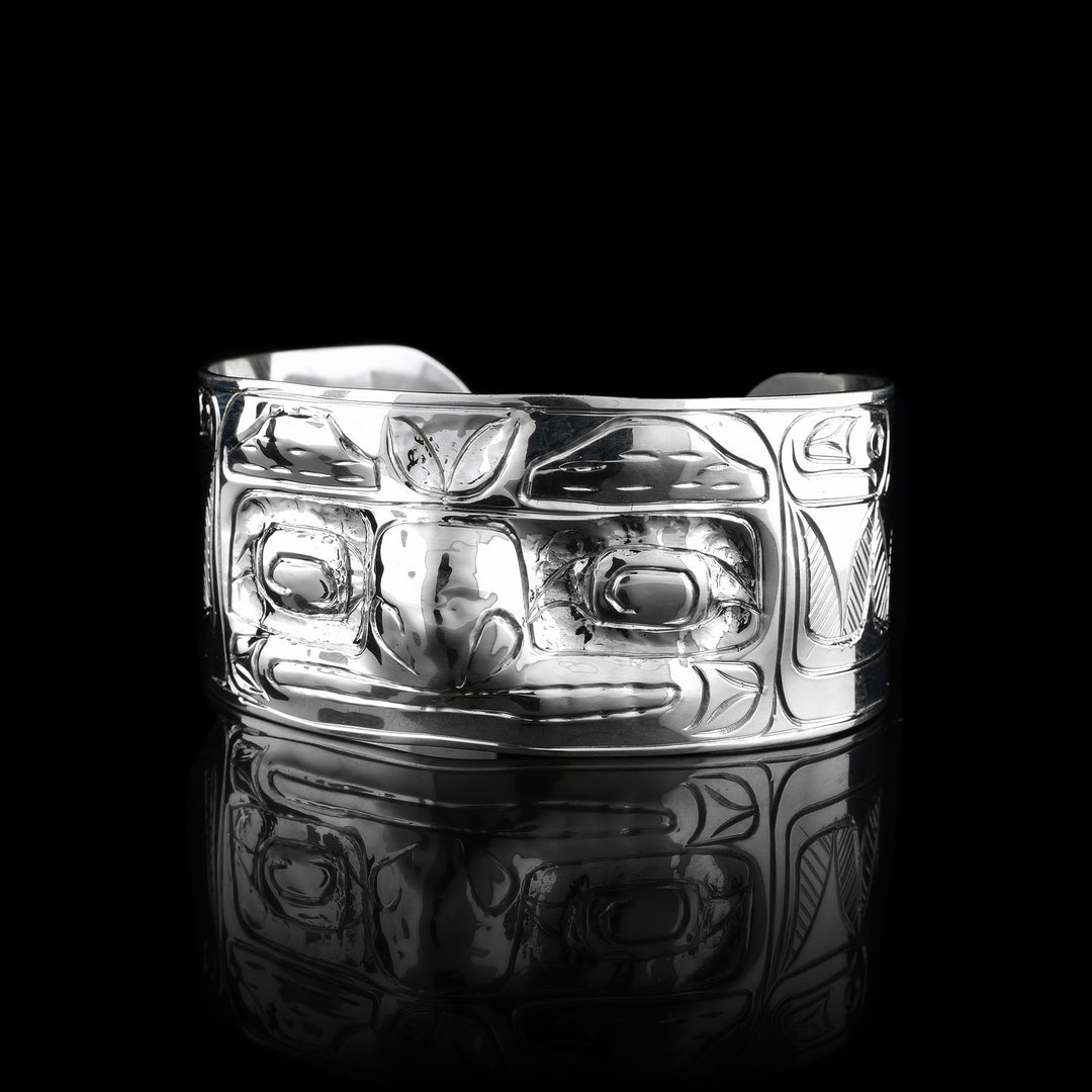 This Raven cuff bracelet has a single, wide band with a gap in the back that is made from sterling silver. There are carvings on the band depicting the face of the Raven legend facing out of the band. The eyes and nose have been shaped into 3D shapes with the eyes going into the piece and the nose coming out. There are feather design to either side of the depiction of the face.