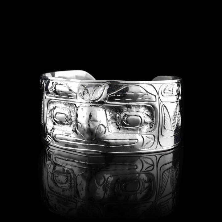 This Raven cuff bracelet has a single, wide band with a gap in the back that is made from sterling silver. There are carvings on the band depicting the face of the Raven legend facing out of the band. The eyes and nose have been shaped into 3D shapes with the eyes going into the piece and the nose coming out. There are feather design to either side of the depiction of the face.