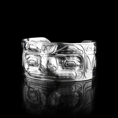 This Raven cuff bracelet has a single, wide band with a gap in the back that is made from sterling silver. There are carvings on the band depicting the face of the Raven legend facing out of the band. The eyes and nose have been shaped into 3D shapes with the eyes going into the piece and the nose coming out. There are feather design to either side of the depiction of the face.