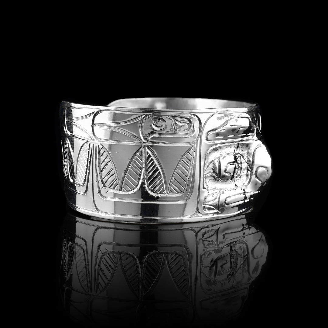 This Raven cuff bracelet has a single, wide band with a gap in the back that is made from sterling silver. There are carvings on the band depicting the face of the Raven legend facing out of the band. The eyes and nose have been shaped into 3D shapes with the eyes going into the piece and the nose coming out. There are feather design to either side of the depiction of the face.