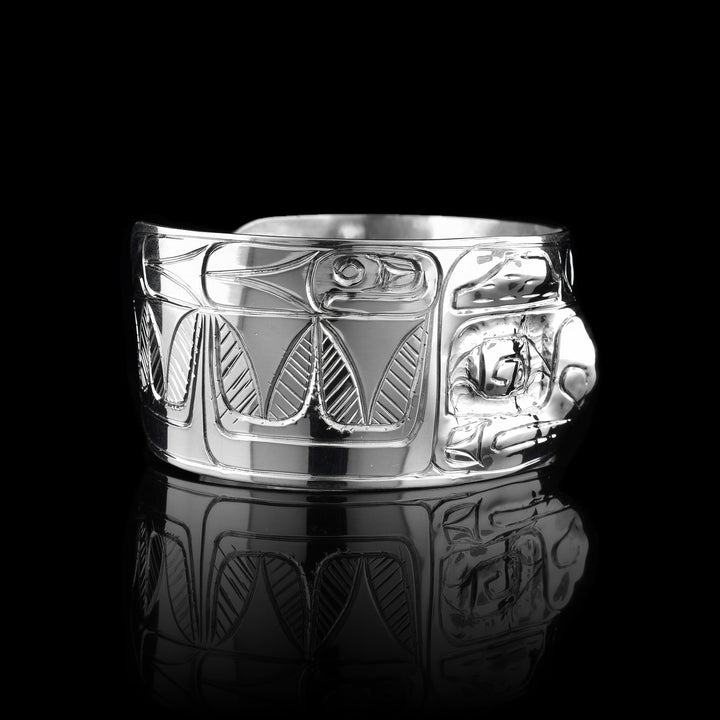 This Raven cuff bracelet has a single, wide band with a gap in the back that is made from sterling silver. There are carvings on the band depicting the face of the Raven legend facing out of the band. The eyes and nose have been shaped into 3D shapes with the eyes going into the piece and the nose coming out. There are feather design to either side of the depiction of the face.