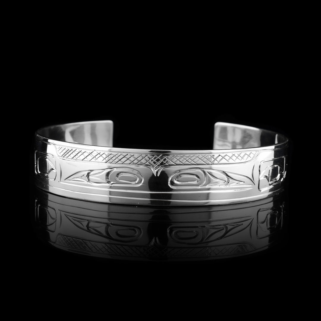 This double Raven bracelet has a single, sterling silver band with a gap In the back. There are two unique depictions of the raven carved into the band. Their heads are shown at the front of the band in a side profile and they are facing away from each other. There are designs on both ends of the band that represent wings.