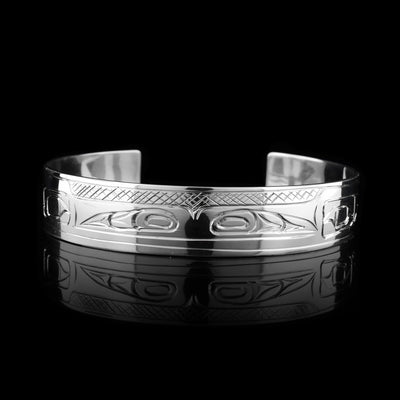 This double Raven bracelet has a single, sterling silver band with a gap In the back. There are two unique depictions of the raven carved into the band. Their heads are shown at the front of the band in a side profile and they are facing away from each other. There are designs on both ends of the band that represent wings.