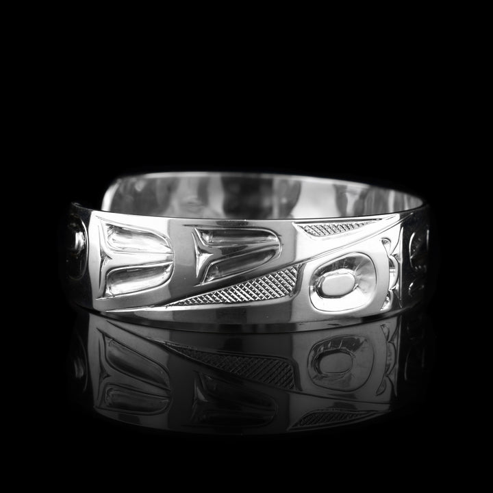 This Eagle bangle bracelet has a single, sterling silver band with a gap in the back. The head of the Eagle is shown in the front at a side profile. The head is large and stretches across most of the band. On either end of the band are wing designs and there are cross hatching designs in the background of the scene.