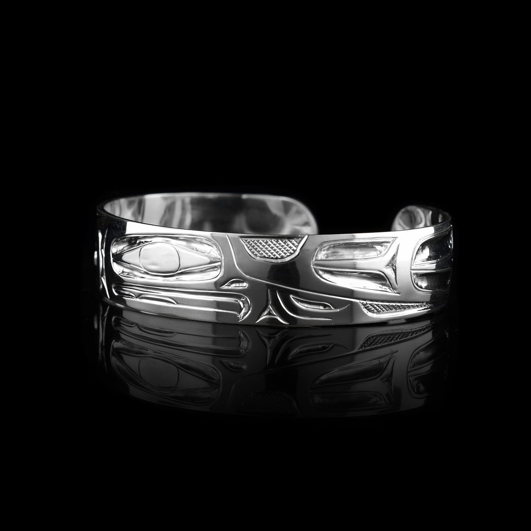 This Eagle bangle bracelet has a single, sterling silver band with a gap in the back. The head of the Eagle is shown in the front at a side profile. The head is large and stretches across most of the band. On either end of the band are wing designs and there are cross hatching designs in the background of the scene.
