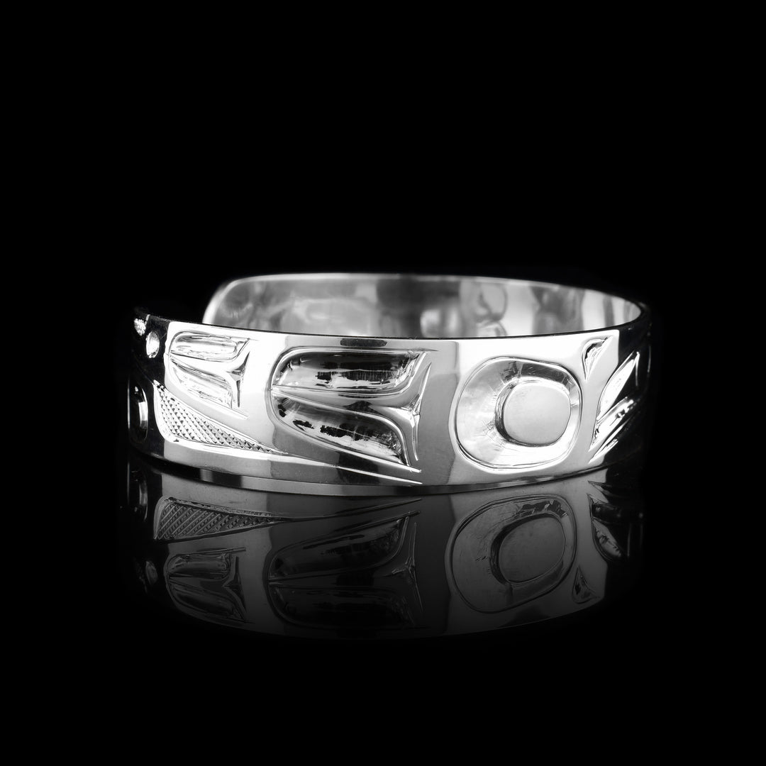 This Raven bangle bracelet has a single, sterling silver band with a gap in the back. The head of the Raven is shown in the front at a side profile. The head is large and stretches across most of the band. On either end of the band are wing designs and there are cross hatching designs in the background of the scene.